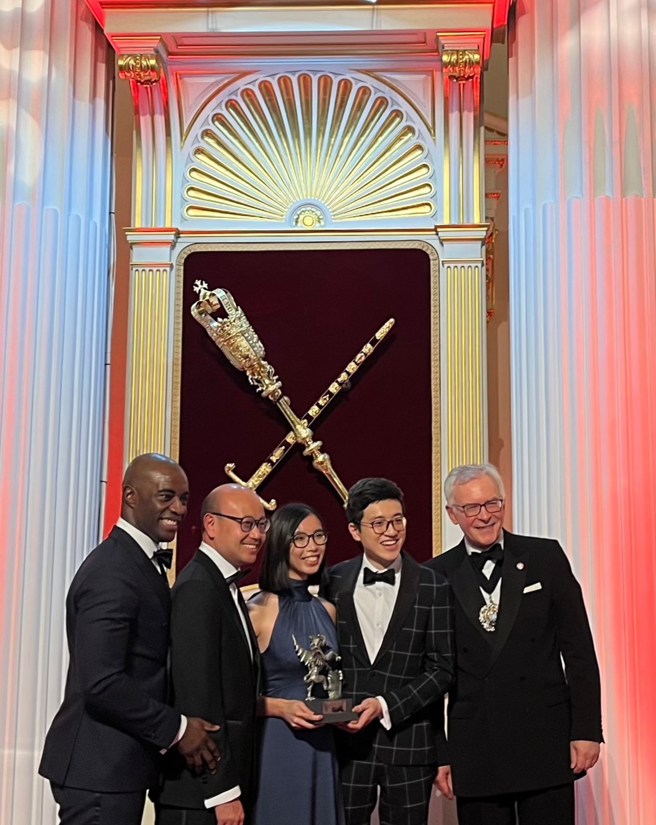 A group of 5 smiling people stand in front of a burgundy background, holding a dragon-shaped award